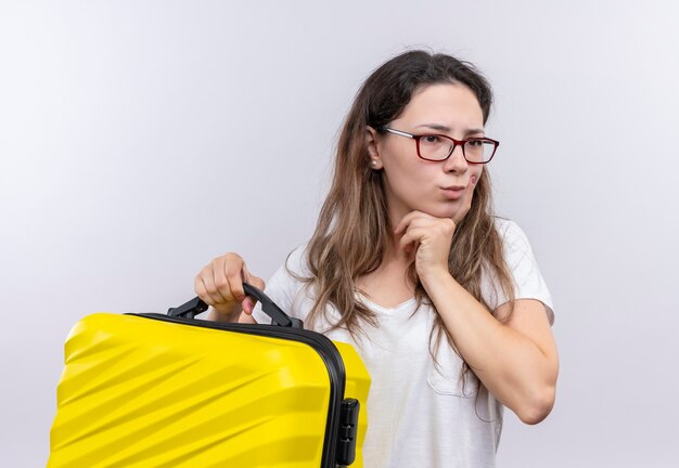 Jovem com uma camiseta branca segurando uma mala de viagem olhando para o lado com uma expressão pensativa no rosto