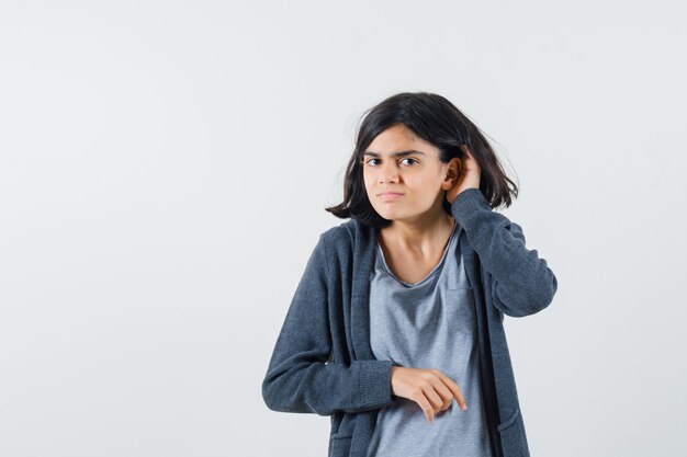 Jovem com uma camiseta cinza claro e um capuz cinza escuro com zíper na frente levantando a mão perto da orelha tentando ouvir algo e parecendo focada