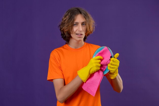 Jovem com uma camiseta laranja usando luvas de borracha, lavando o prato, olhando para a câmera com um sorriso confiante em pé sobre um fundo roxo