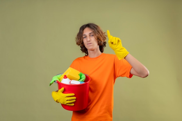 Jovem com uma camiseta laranja usando luvas de borracha segurando um balde com ferramentas de limpeza apontando para cima tendo uma ótima ideia, sorrindo confiante em pé sobre um fundo verde
