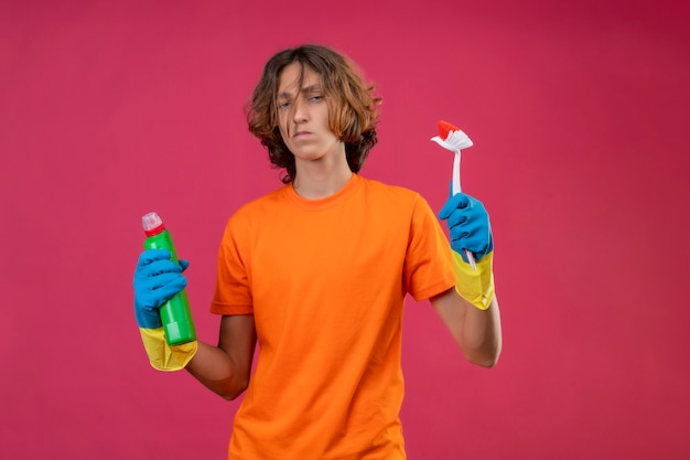 Jovem com uma camiseta laranja usando luvas de borracha segurando um frasco de material de limpeza e uma escova olhando para a câmera descontente em pé sobre um fundo rosa