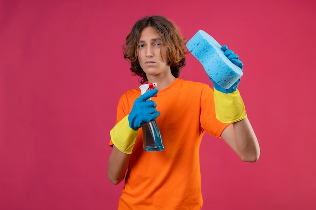 Jovem com uma camiseta laranja usando luvas de borracha, segurando um spray de limpeza e uma esponja, parecendo confuso em pé sobre um fundo rosa