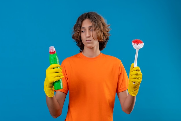 Jovem com uma camiseta laranja usando luvas de borracha segurando uma escova e um frasco com material de limpeza parecendo incerto, tendo dúvidas em pé sobre um fundo azul