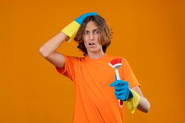 Jovem com uma camiseta laranja usando luvas de borracha segurando uma escova olhando para ela surpreso tocando a cabeça em pé sobre o fundo amarelo