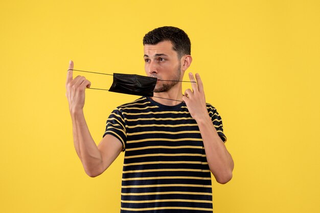 Jovem com uma camiseta listrada em preto e branco segurando uma máscara preta em fundo amarelo