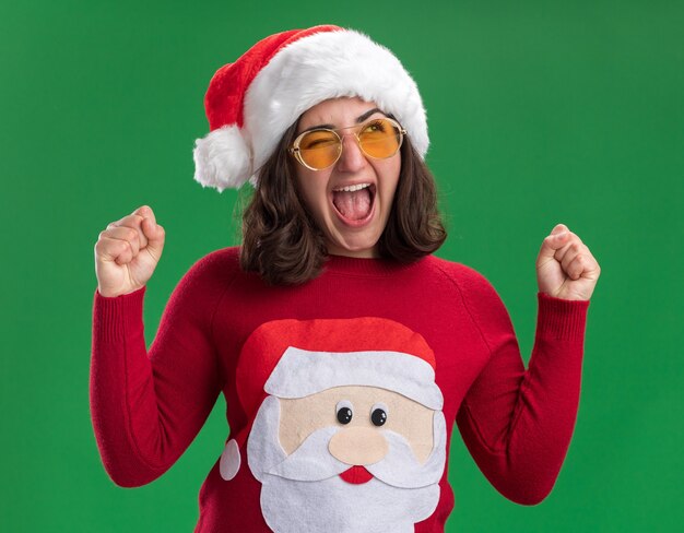 Foto grátis jovem com uma camisola de natal com chapéu de papai noel e óculos, gritando animada e louca feliz cerrando os punhos em pé sobre a parede verde