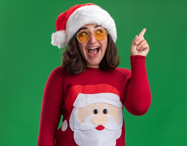 Jovem com uma camisola de Natal com chapéu de Papai Noel e óculos, olhando feliz e alegre apontando com o dedo indicador em pé sobre a parede verde