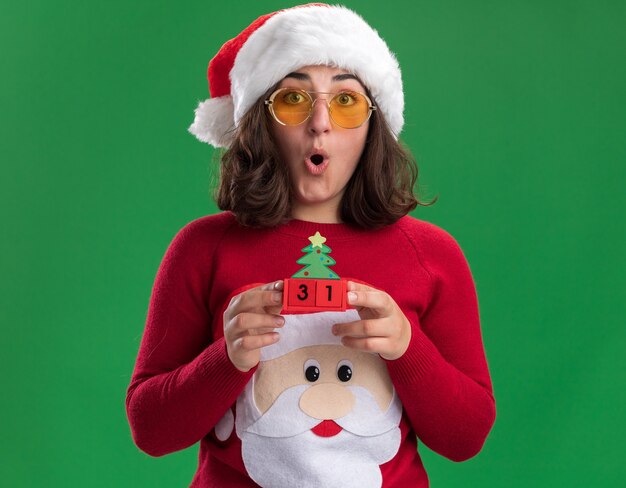 Jovem com uma camisola de Natal com chapéu de Papai Noel e óculos segurando cubos de brinquedo com data de ano novo espantada e surpresa em pé sobre a parede verde