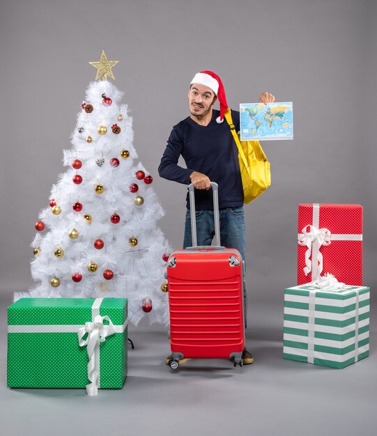 Jovem com uma mochila amarela segurando um mapa e uma valise perto da árvore de natal e presentes em cinza