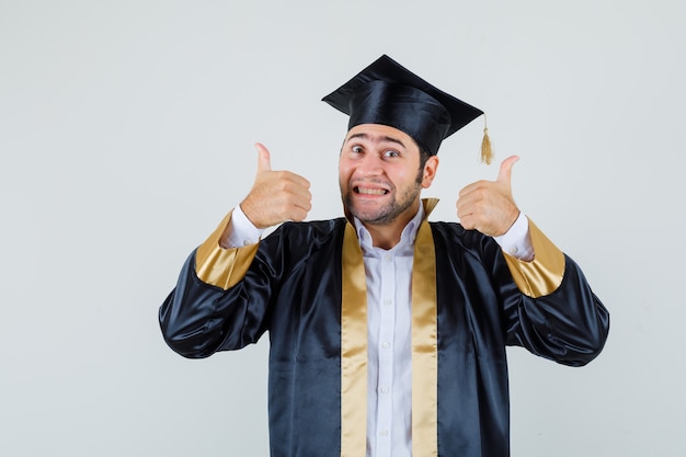 Jovem com uniforme de pós-graduação, mostrando os polegares duplos e olhando alegre, vista frontal.