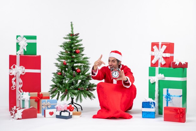 Jovem comemora o feriado de natal sentado no chão e mostrando o relógio perto de presentes e uma árvore de natal decorada