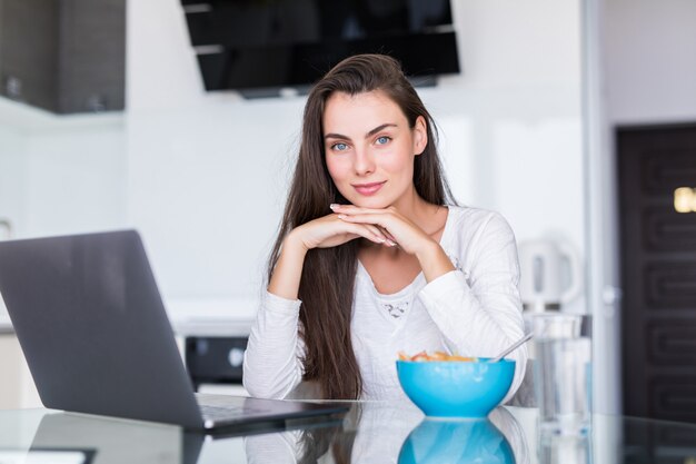 Jovem comer salada trabalhando no laptop na cozinha