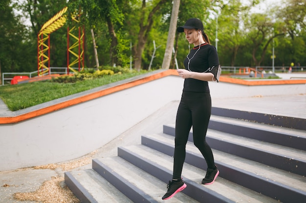 Jovem concentrada atlética linda mulher de uniforme preto, boné com fones de ouvido, fazendo exercícios esportivos, aquecimento antes de descer correndo as escadas no parque da cidade ao ar livre