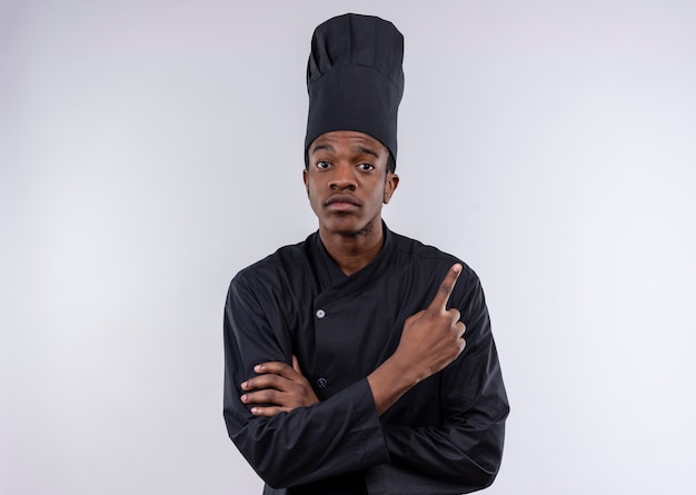 Jovem confiante afro-americana com uniforme de chef cruza os braços e aponta para cima, isolada no fundo branco com espaço de cópia