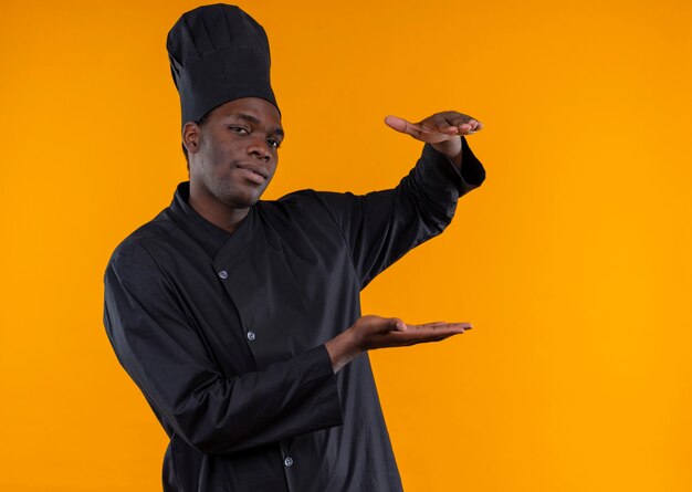 Jovem confiante afro-americana com uniforme de chef fingindo segurar algo isolado em um fundo laranja com espaço de cópia