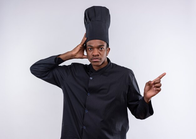Jovem confiante afro-americana com uniforme de chef segura a cabeça e aponta para o lado isolado no fundo branco com espaço de cópia