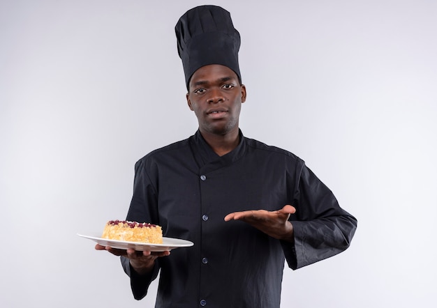 Jovem confiante afro-americana com uniforme de chef segura e aponta para o bolo no prato branco com espaço de cópia