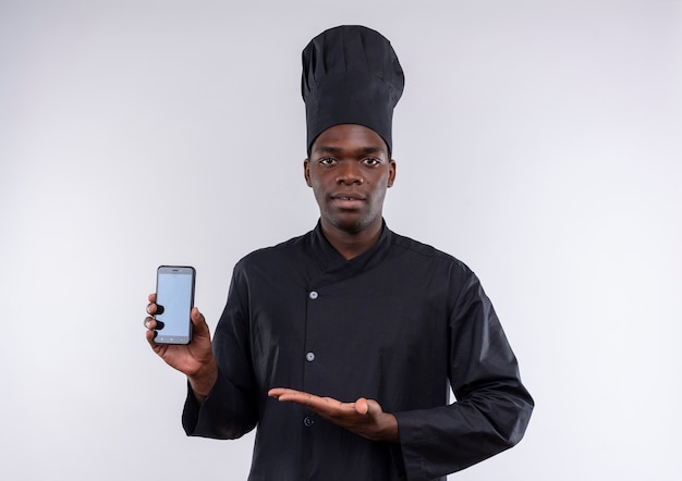 Jovem confiante afro-americana com uniforme de chef segura e aponta para o telefone em branco com espaço de cópia