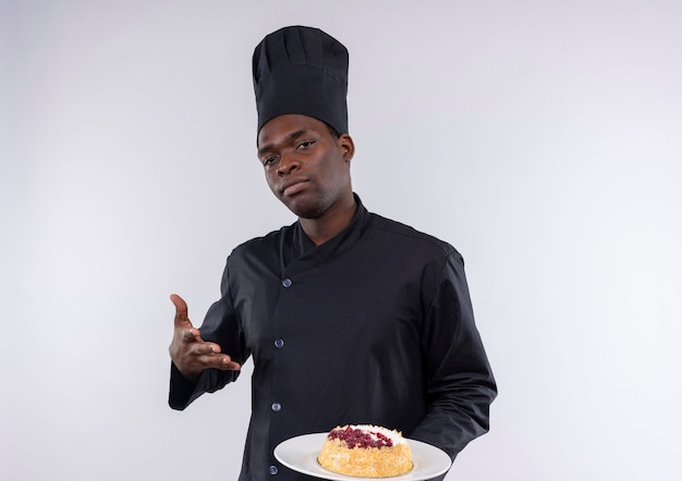 Jovem confiante afro-americana com uniforme de chef segura e aponta para um bolo no prato, olhando para a câmera em branco com espaço de cópia