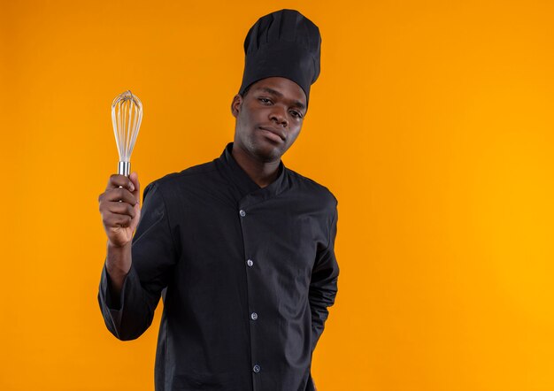 Jovem confiante afro-americana com uniforme de chef segurando um batedor olhando para a câmera isolada em um fundo laranja com espaço de cópia