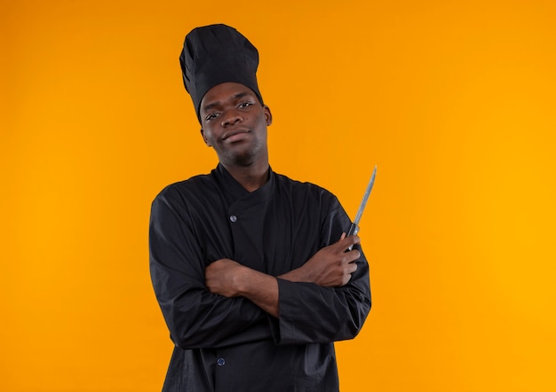 Jovem confiante afro-americana com uniforme de chef segurando uma faca com os braços cruzados, isolada em um fundo laranja com espaço de cópia