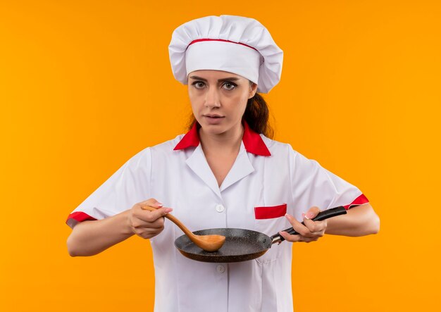 Jovem confiante caucasiana cozinheira com uniforme de chef segurando uma frigideira e uma colher de madeira isoladas na parede laranja com espaço de cópia