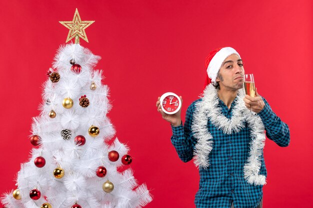 Jovem confiante com chapéu de papai noel e degustando uma taça de vinho e um relógio em pé perto da árvore de natal