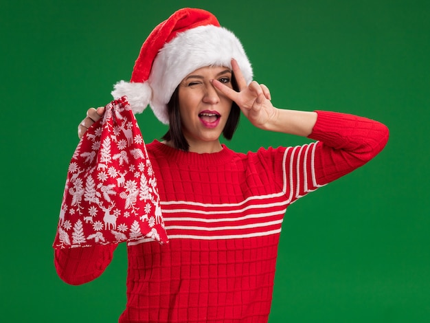 Jovem confiante com chapéu de Papai Noel segurando um saco de presente de Natal, olhando para a câmera piscando, mostrando o símbolo do sinal-V perto do olho isolado no fundo verde