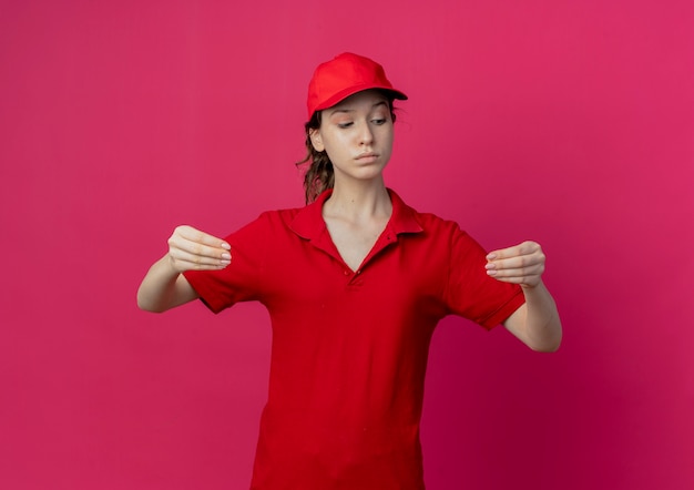 Jovem confiante e bonita entregadora de uniforme vermelho e boné olhando para baixo e fingir que está segurando algo isolado em um fundo carmesim