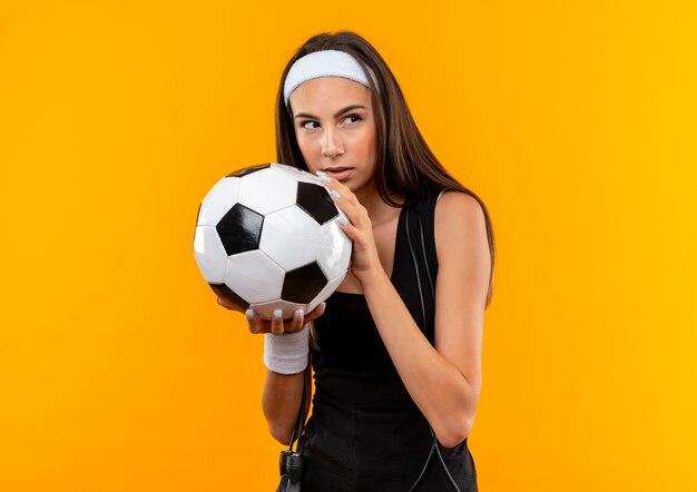 Jovem confiante e bonita esportiva usando bandana e pulseira, segurando uma bola de futebol, olhando para o lado com uma corda de pular em volta do pescoço isolada na parede laranja