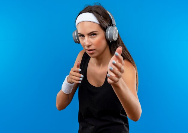 Jovem confiante e bonita esportiva usando bandana, pulseira e fones de ouvido, estendendo as mãos isoladas na parede azul