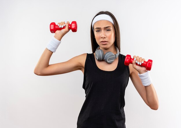 Jovem confiante e bonita esportiva usando bandana, pulseira e fones de ouvido no pescoço, levantando halteres isolados na parede branca