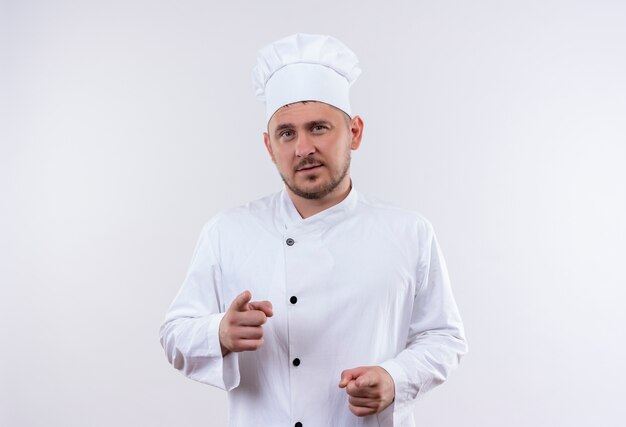 Jovem confiante e bonito cozinheiro com uniforme de chef apontando isolado na parede branca