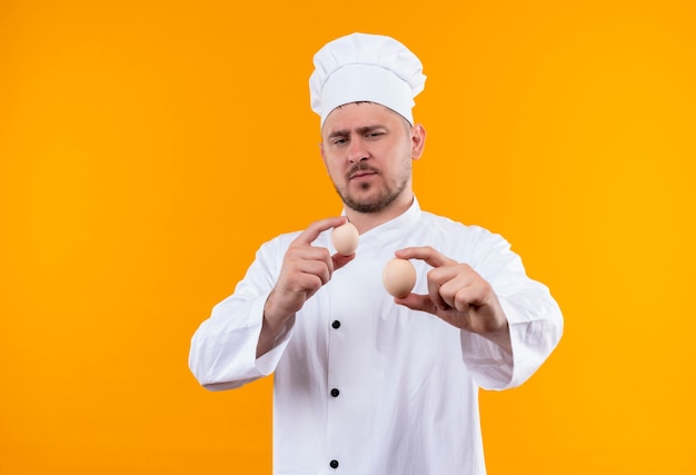 Jovem confiante e bonito cozinheiro em uniforme de chef segurando e olhando para ovos isolados na parede laranja