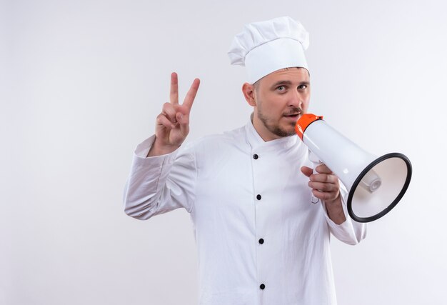 Jovem confiante e bonito cozinheiro em uniforme de chef, segurando o alto-falante e fazendo o sinal da paz, isolado na parede branca