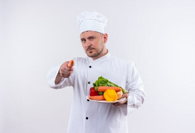 Jovem confiante e bonito cozinheiro em uniforme de chef, segurando o prato com legumes e apontando com cenoura na parede branca isolada