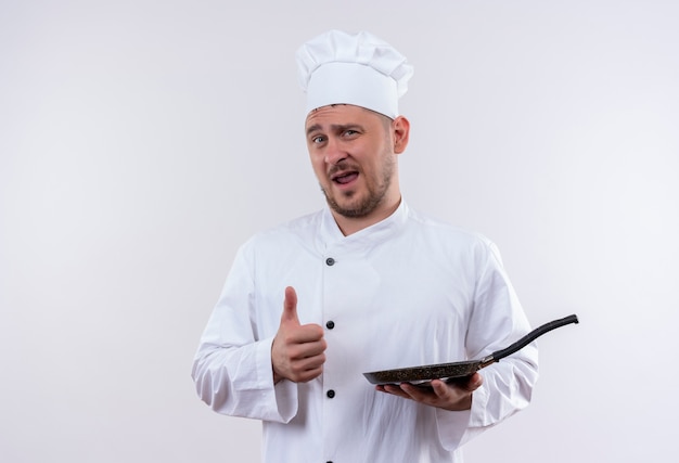 Jovem confiante e bonito cozinheiro em uniforme de chef segurando uma frigideira aparecendo o polegar isolado na parede branca