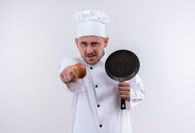 Jovem confiante e bonito cozinheiro em uniforme de chef segurando uma frigideira e apontando com uma colher na parede branca isolada