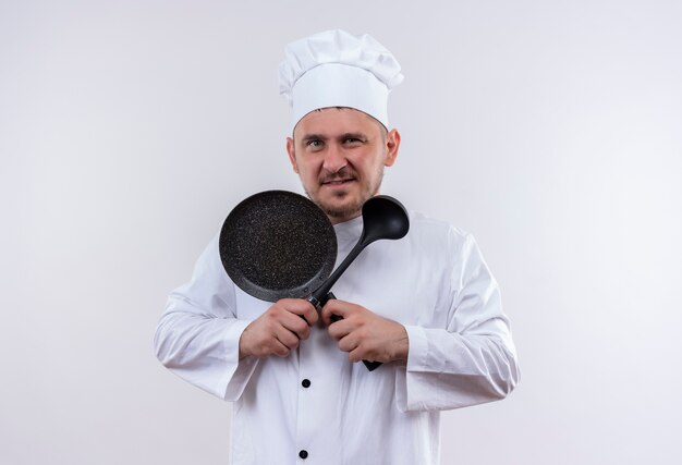 Jovem confiante e bonito cozinheiro em uniforme de chef segurando uma frigideira e uma concha isoladas na parede branca