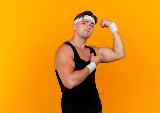 Jovem confiante e bonito homem desportivo usando bandana e pulseiras, gesticulando com força e apontando para os músculos isolados em laranja com espaço de cópia