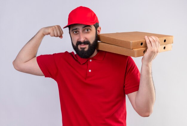 Jovem confiante e bonito homem entregador caucasiano vestindo uniforme vermelho e boné segurando caixas de pizza no ombro e gesticulando forte isolado no branco