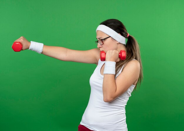 Jovem confiante e desportiva com óculos ópticos, bandana e pulseira segurando halteres