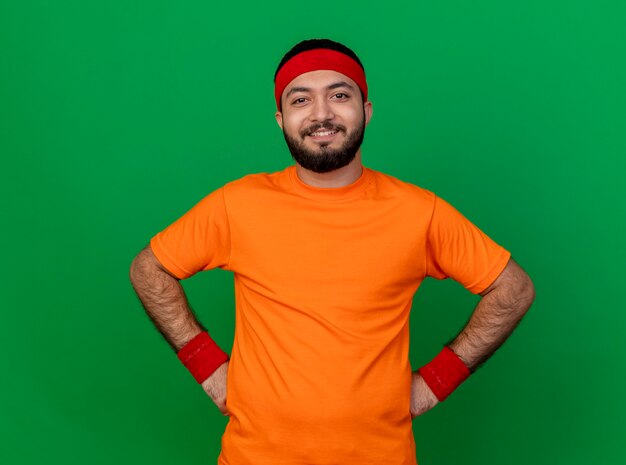 Foto grátis jovem confiante e esportivo usando bandana e pulseira, colocando as mãos no quadril, isolado sobre fundo verde