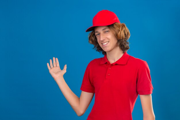 Jovem confiante entrega uniforme vermelho sorrindo amigável apresentando algo com mão sobre parede azul isolada