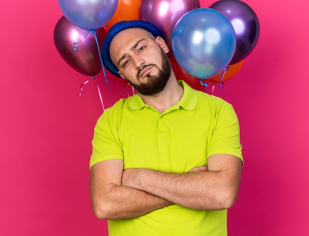 Jovem confiante inclinando a cabeça usando um chapéu de festa azul em pé na frente de balões, cruzando as mãos isoladas na parede rosa