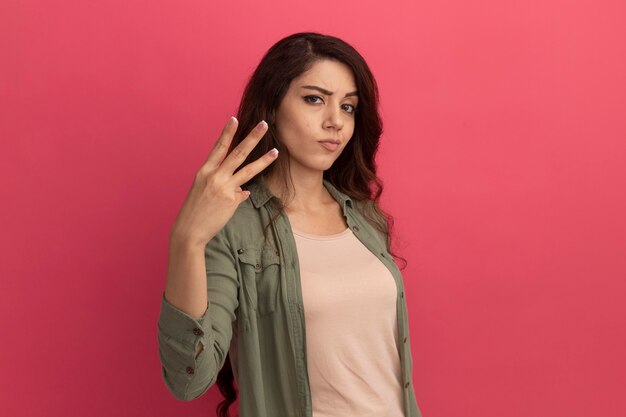 Jovem confiante linda com uma camiseta verde oliva mostrando três isolados na parede rosa