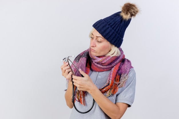 Foto grátis jovem confiante loira doente com chapéu de inverno e cachecol segura e olha para o estetoscópio isolado na parede branca com espaço de cópia