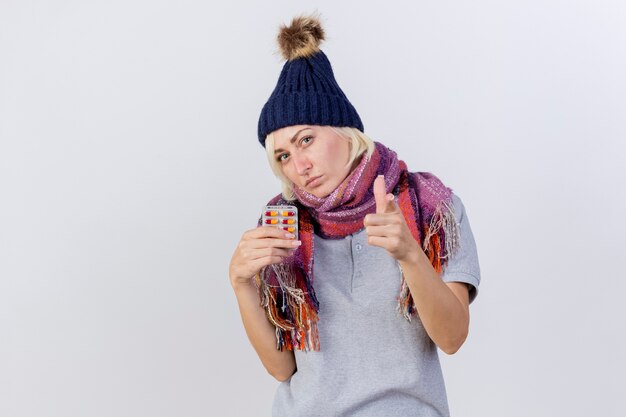 Jovem confiante loira doente, mulher eslava usando chapéu e lenço de inverno segurando um pacote de pílulas médicas e pontos isolados na parede branca com espaço de cópia