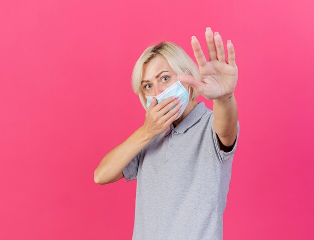 Foto grátis jovem confiante loira doente, mulher eslava usando máscara médica, coloca a mão na boca e fica de pé com a mão levantada, isolada na parede rosa com espaço de cópia