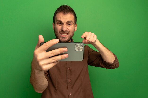 Jovem confiante segurando e olhando para o celular, apontando para ele isolado na parede verde